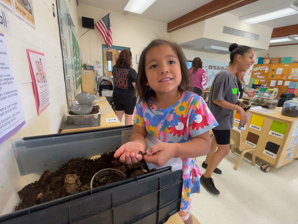 Young girl in a floral dress holds soil in a classroom, with adults nearby, an American flag, and color labels yellow, green, brown, black.