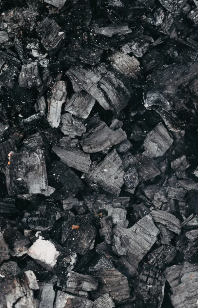Close-up of a pile of black charcoal pieces with rough textures. The dark surface shows some light gray ash, creating a rustic feel.
