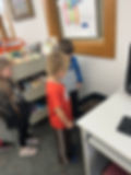 Three children stand in a classroom library beside a book cart and desk, looking at a composter; colorful poster on wall.