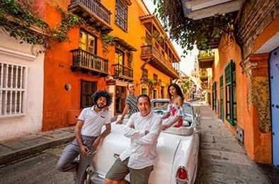 Turisták egy klasszikus nyitott kabrió autóval pózolnak színes, balkonos házakkal szegélyezett óváro