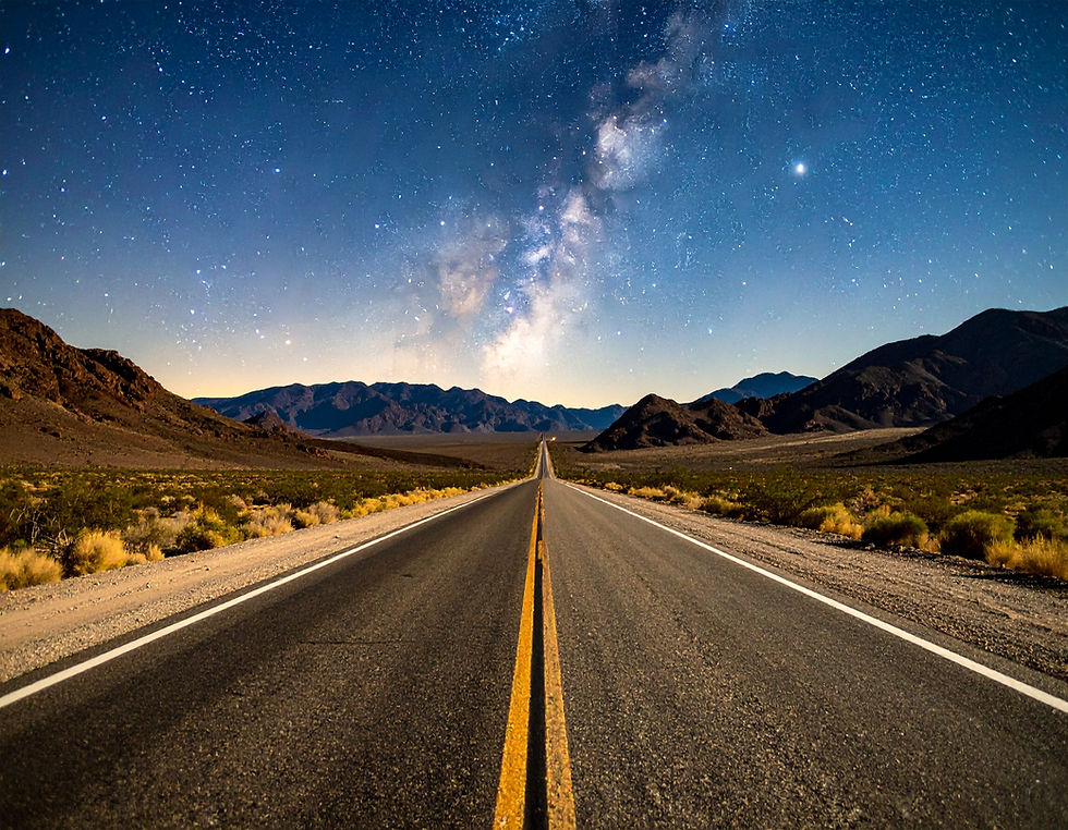 Straight road leading into distant mountains under a starry night sky. Milky Way visible. Desert landscape with a serene, vast feeling.