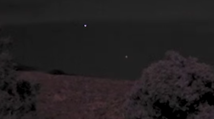 Night sky with two bright dots above a dark landscape; a blurry bush is in the foreground, creating an eerie, mysterious mood.
