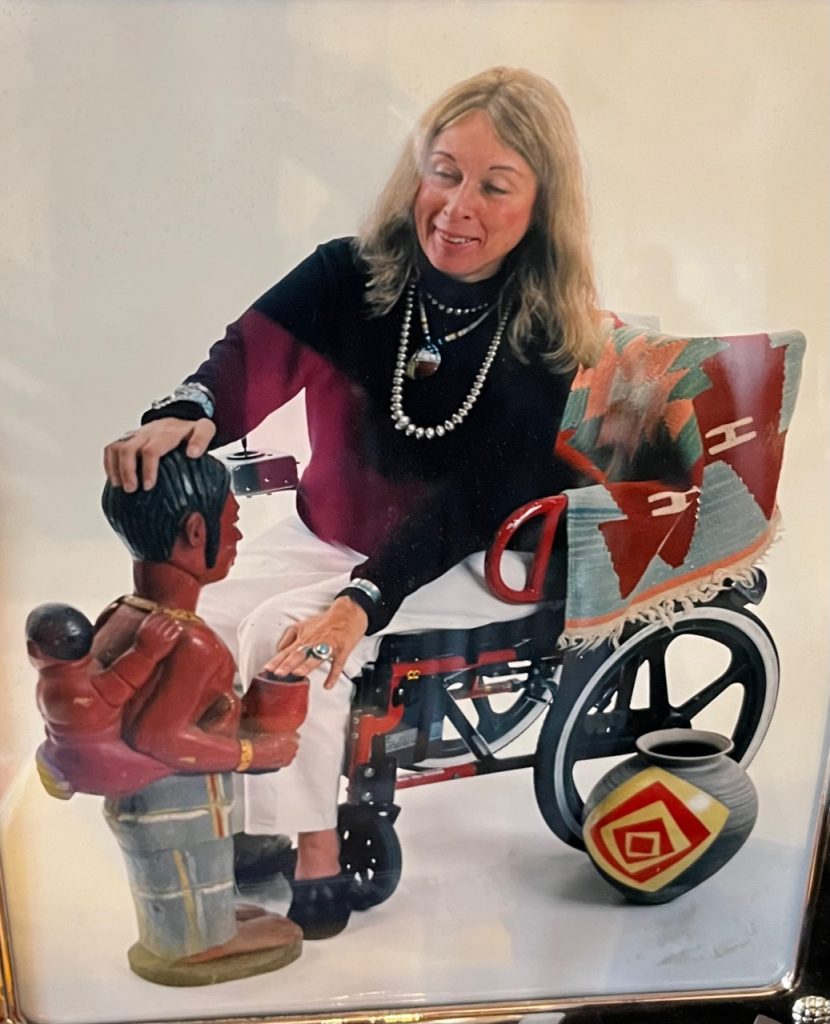 Anita Silvers is depicted. She is sitting in her powerchair, which has snazzy black and red detailing. She has pale skin, long blonde hair that is greying, is wearing white slacks and a black turtleneck, and wearing large beaded silver jewelry including necklaces and a turqoise ring. She has a blanket across the back of her chair colorfully decorated in geometric patterns common in North American indigenous peoples' blankets. She is gazing at and touching a ceramic sculpture of a mesoamerican person wearing an infant on their back. A ceramic pot rests on the floor next to the left wheel of her chair.