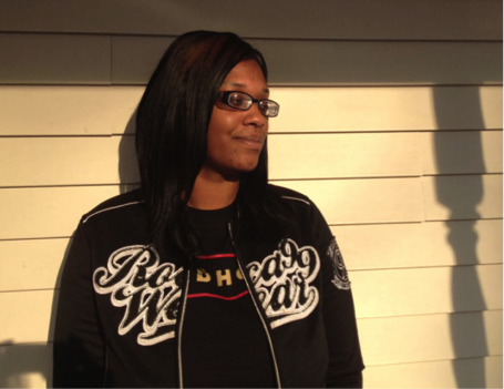 Tamesha Means is a young African-American woman with long, straight black hair. She is wearing rectangular glasses, a black t-shirt, and a lightweight black jacket. She leans against the aluminum-sided wall of a house, the shadows cast by a porch roof falling just above her head. She is bathed in sunlight. She has a small, but not happy, smile on her face. IMAGE CREDIT: ACLU