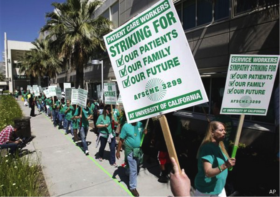 IMAGE DESCRIPTION: A long line of men and women with various skin tones in green t-shirts walks a line chalked on a sidewalk, disappearing into the distance. Each person holds a picket sign in green and white. Signs read “Patient Care Workers Striking for: Our patients, Our family, Our future AFSCME 3299 at University of California” and “Service Workers Sympathy Striking for: Our patients, Our family, Our future AFSCME 3299 at University of California.” The latter in particular shows solidarity where non-health care providers lend their work actions to the cause of others. The image pertains to a May 21, 2013 work action in front of UC Davis Medical Center in Sacramento, CA, in the U.S. IMAGE CREDIT: AP