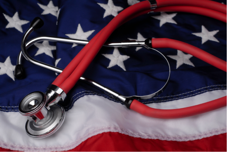 A healthcare provider’s stethoscope, its flexible tubing colored a bright red, lays atop an American flag with its red and white stripes, and blue field covered in white stars. 