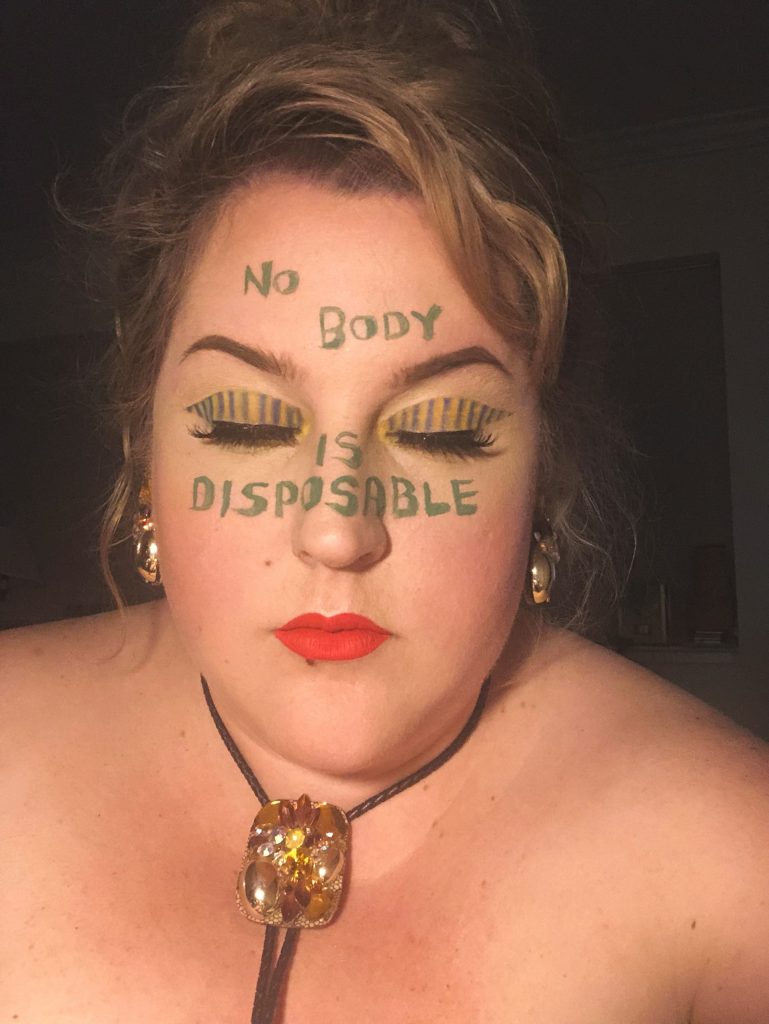 A fat woman with pale skin wearing a braided leather necklace with a decorative glass fastener is seen close-up with her eyes closed. On her eyelids is an elaborate pattern of vertical stripes. On her forehead is written "NO BODY" and across her nose and cheeks is written "IS DISPOSABLE." 