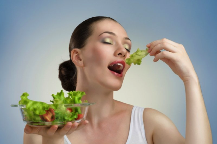 A Caucasian woman with dark hair slicked back into a bun and elaborate makeup, wearing a clean white tank top, is holding a salad which is just green lettuce and red sliced tomatoes. She is not eating it with a fork and it appears to have no dressing or embellishment. She is holding a piece of lettuce above her mouth with her head tilted slightly back, eyes closed ecstatically. This must be the most amazing salad ever. This image illustrates a trend in such images that many folks call “women eating salad.” It is cross-cultural.