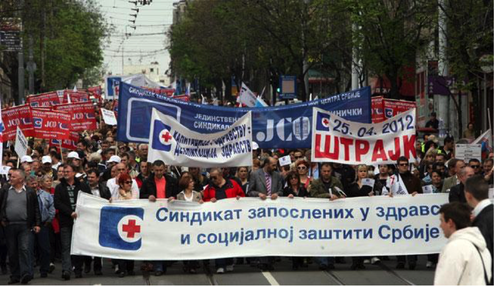 IMAGE DESCRIPTION: In April of 2012, trade unions of health and social protection workers in Belgrade, Serbia, organized a one-hour “warning strike” in response to a state proposal to reduce the salaries of all healthcare employees by 10 percent. In this image, a dense crowd or professionally dressed individuals marches down a major tree-lined street. All the people who can be seen are light-skinned; both men and women are present. Street car tracks can be seen in the road surface disappearing under the feet of the crowd. Signs are not legible to this author as they are written in Cyrillic alphabet. However, the very large banners and small signs alike bear a common symbol: a blue square with rounded corners; in the middle of the right side of the square is a white circle within which is a red cross. The red cross and the color blue are traditional international symbols of modern medicine. Some signs bear the date “April 25, 2012” in European format: 25.4.12. At the bottom of this blog entry, you can find a description of more recent strikes in Serbia over similar austerity measures, as well as other examples of health care provider work actions in Asia, Europe, Africa, and North America. IMAGE CREDIT: b92.