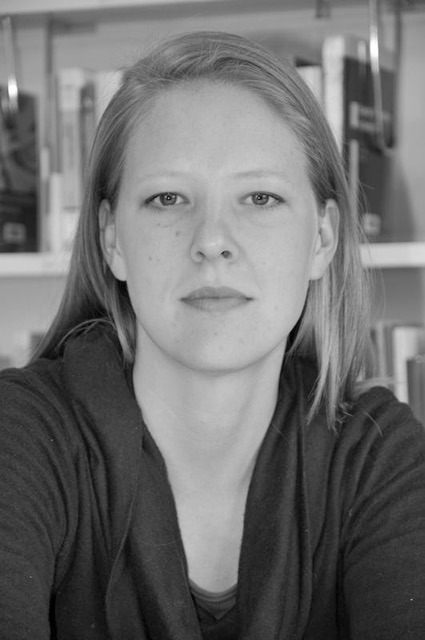 This image shows a woman with pale skin and long blonde hair. A bookshelf is behind her as though the picture is taken in a professional office. She stares seriously at the photographer.