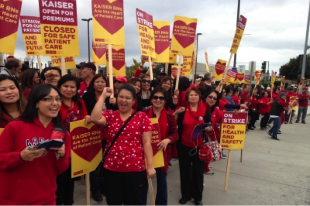 IMAGE DESCRIPTION: A crowd of mostly women and some men wears red shirts and sweaters. Most hold picket signs (large paper bearing messages stapled to wooden pickets). The people are smiling at the camera. The line of picketers extends into the distance. Signs are read and yellow and bear messages such as “On Strike for Health and Safety”, “Kaiser open for premiums Close for safe patient care”, and “Striking for our patients and our future”. A caption provided by the image source indicates that these nurses were at Kaiser Vallejo in California, and that they are protesting lack of supplies and training as well as short staffing, issues that affect Ebola preparedness but also more common safety risks. IMAGE CREDIT: California Nurses Association