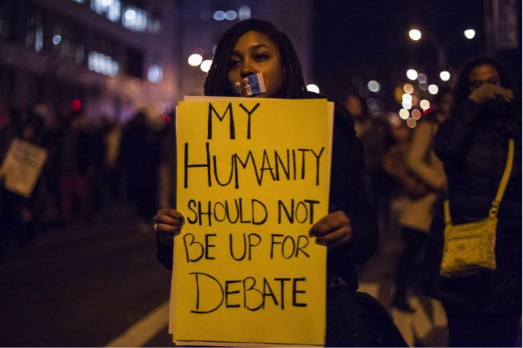 A young black woman, her shoulder length hair in medium-thickness dredlocks, is in the streets of a major city. Her mouth is covered with a piece of red, white, and blue-striped tape on which are written the words “I can’t breathe,” the last words spoken by Eric Garner. There are many people walking in the street behind her, some black and some white, though it is hard to tell as the image is out of focus behind her. She carries a bright yellow sign which says in bold, black, capital letter, “My humanity should not be up for debate.” Karla Holloway writes about the way that people who move through the world in black bodies have their private personhood affected by the public text of their bodies. IMAGE CREDIT: REUTERS/Elizabeth Shafiroff