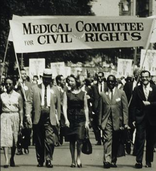 This black and white photo from the 1960's shows a group of both white and black people in professional dress marching under a banner which reads "Medical Committee for Civil Rights." Behind them can be scene marchers receding into the distance, many carrying posters showing the symbol of medicine, the rod of Aesclepius or staff of Hermes, a staff with two winged snakes curled around it. At the bottom of those signs are the letters MD, for medical doctor.