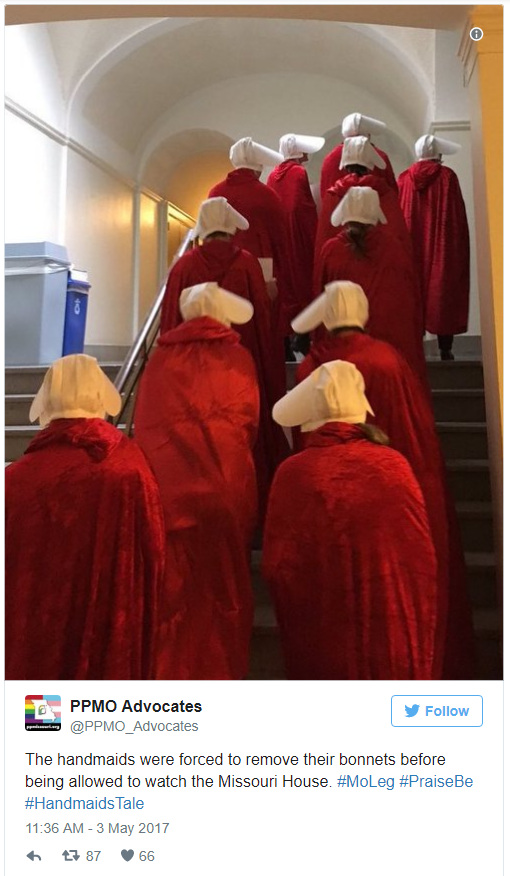 Walking 2 by 2, women in red cloaks with white bonnets, like the costumes worn by Handmaids in the Hulu series, ascend a staircase to enter the chambers of the Missouri Legislature to watch the House debate a bill in May. The picture was posted on the Twitter account of Planned Parenthood Missouri Advocates. The text reads "The handmaids were forced to remove their bonnets before being allowed to watch the Missouri House. #MoLeg #PraiseBe #HandmaidsTale." It is dated May 3 of 2017.