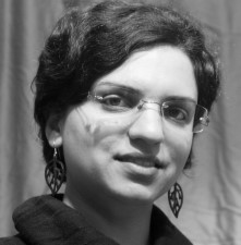 A black and white photo of a woman with short wavy dark hair. She is wearing earings shaped like leaves and a pair of rectangular wire framed glasses. Her mouth is slightly open in a very small smile as though she is listening and about to speak.
