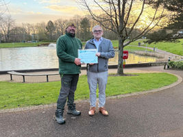 Grounds maintenance operative receiving GEM award from Managing Director in front of landscaped grounds with large pond feature and trees