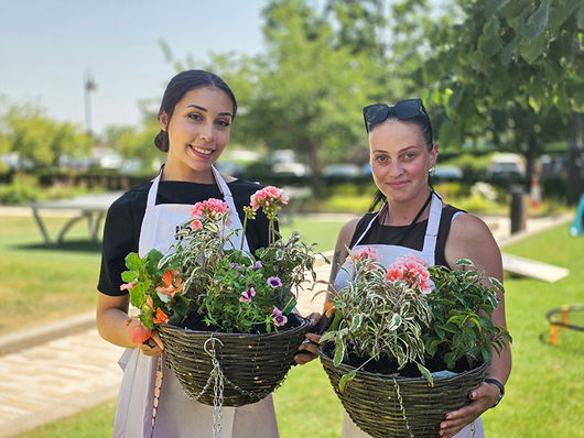 Two women holding hanging baskets with flowers in a garden
