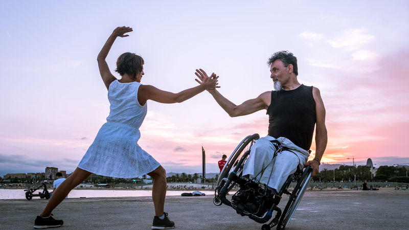 Donna con vestito bianco in piedi che tende la mano ad un uomo in sedia a rotelle in equilibrio su una sola ruota