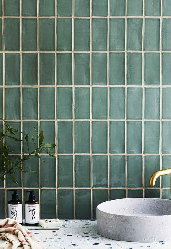 A green glass vertical subway tile backsplash sits behind a terrazzo vanity and with a ceramic basin and a brass tap on top.