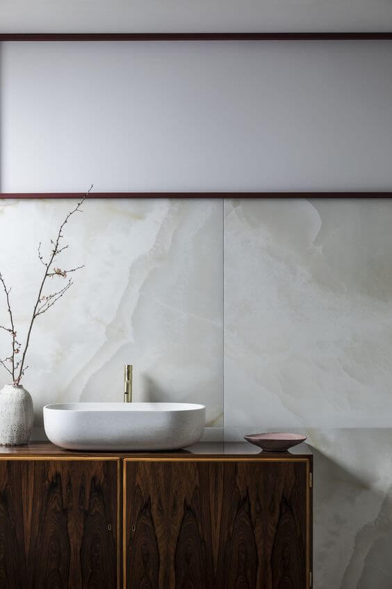 Large porcelain tiles make a backsplash behind a timber vanity with a basin and tap on top. A white vase sits next to the basin holding skinny branches.