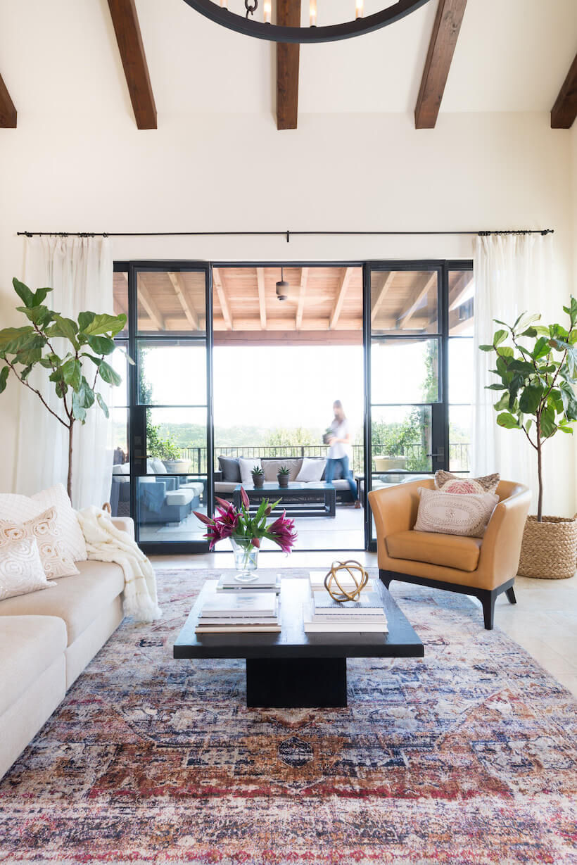 Spanish revival style living room. Exposed dark timber ceiling beams. Large steel doors opening to an outdoor patio with a woman walking by. Two fiddle leaf plants and white drapes flank the doors. Camel-beige leather chair faces a white sofa with white cushions and throw. A dark timber rectangle coffee table sits on top of a deep pink and blue persian style rug.