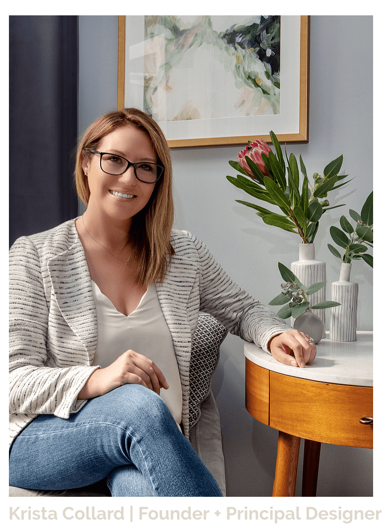 A millennial woman, residential interior designer based on the Lower North Shore of Sydney, Australia. She is wearing a white and grey tweed blazer with a white cami, jeans and black framed glasses. Her hair is dark blonde and she is smiling with her legs crossed and her arm rested on a marble and walnut side table surrounded by native Australian flowers and plants. A watercolour abstract painting in a matted and brass frame hangs on the wall behind her as do charcoal drapes.