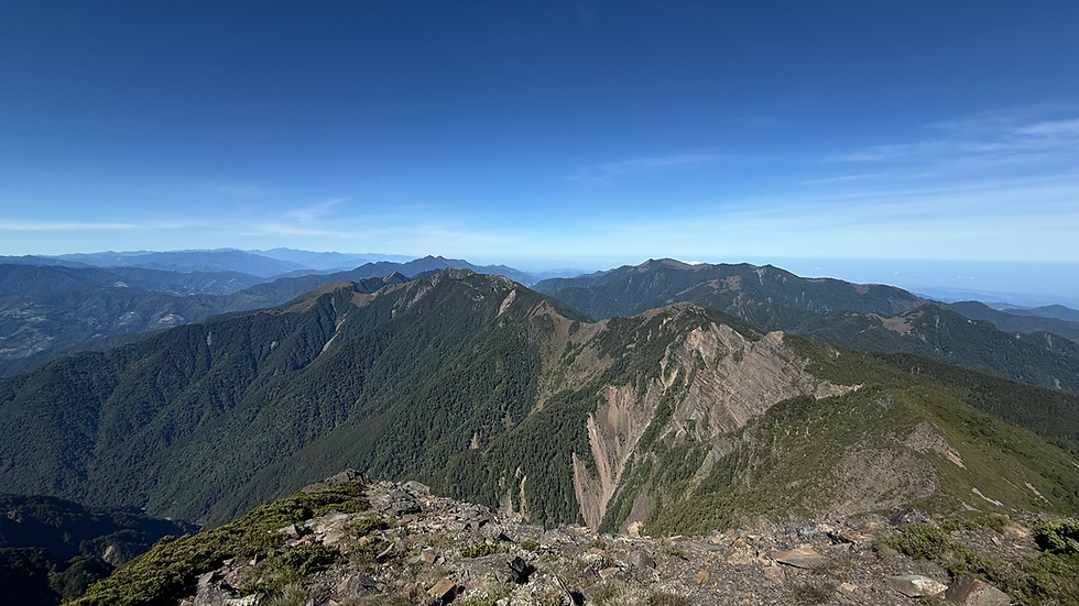 Wide mountain ridge panorama under clear blue sky, with green slopes, rocky peaks, and distant layered mountains fading in haze
