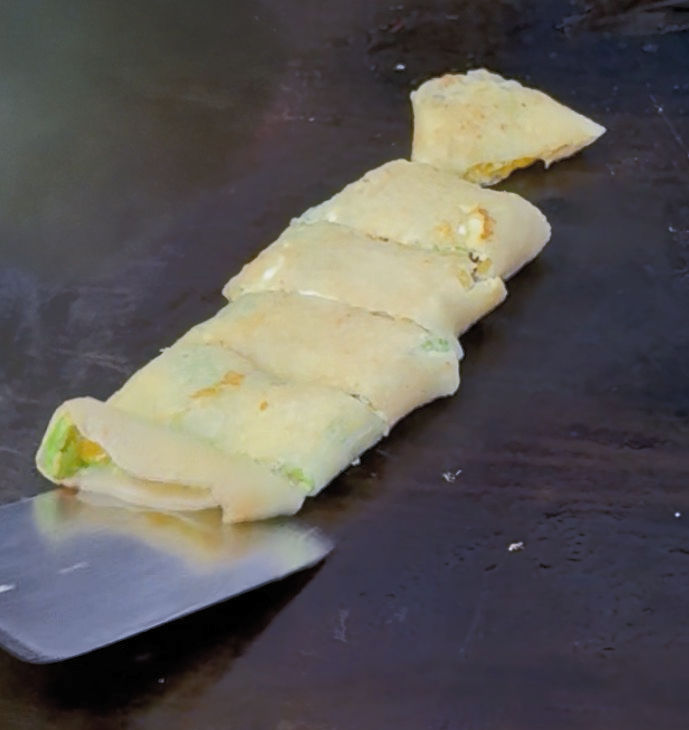 Sliced rolled crepe or omelet on a hot griddle, with a metal spatula lifting one end; pale golden, simple cooking scene