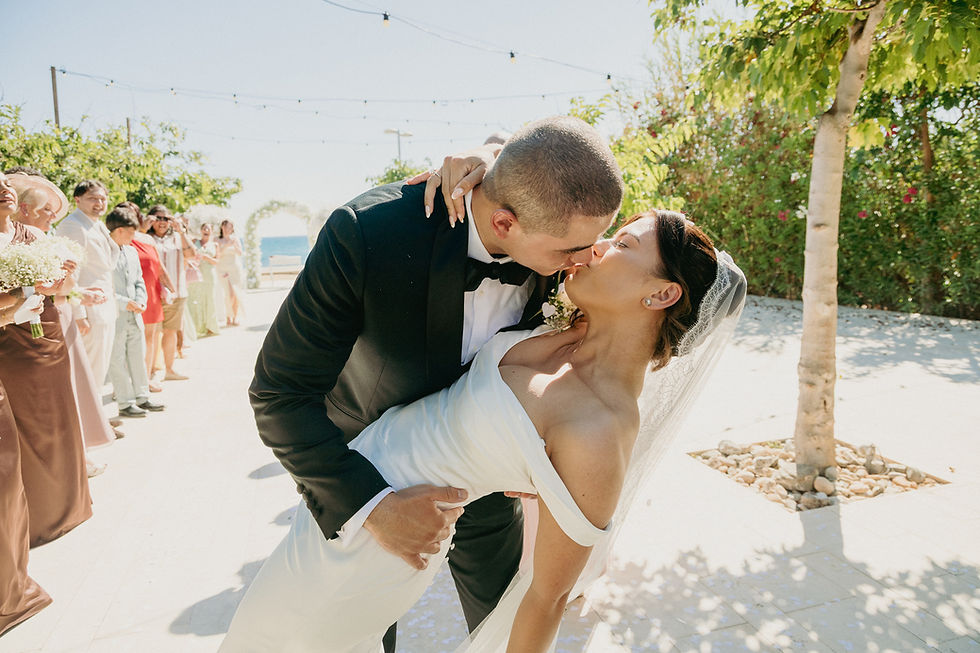 Bride in white dress and groom in black suit kiss during sunny outdoor wedding, with guests lined up and string lights overhead. A Cyprus fairytale wedding