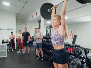 A woman at Peak Fitness and Motion lifting a weight over her head while onlookers are impressed
