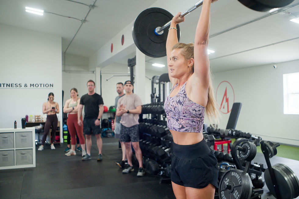 Woman lifting weights at Peak Fitnees and Motion in Bozeman, wearing a floral tank and black shorts. Group of people watching. "Fitness & Motion" visible.