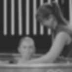 A smiling young white woman baptizes a young white girl. The girl holds her nose & squints her eyes as she comes out of the water, baptized by immersion.