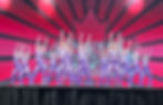 Teenage dance troupe in sparkling purple jumpsuits on stage.