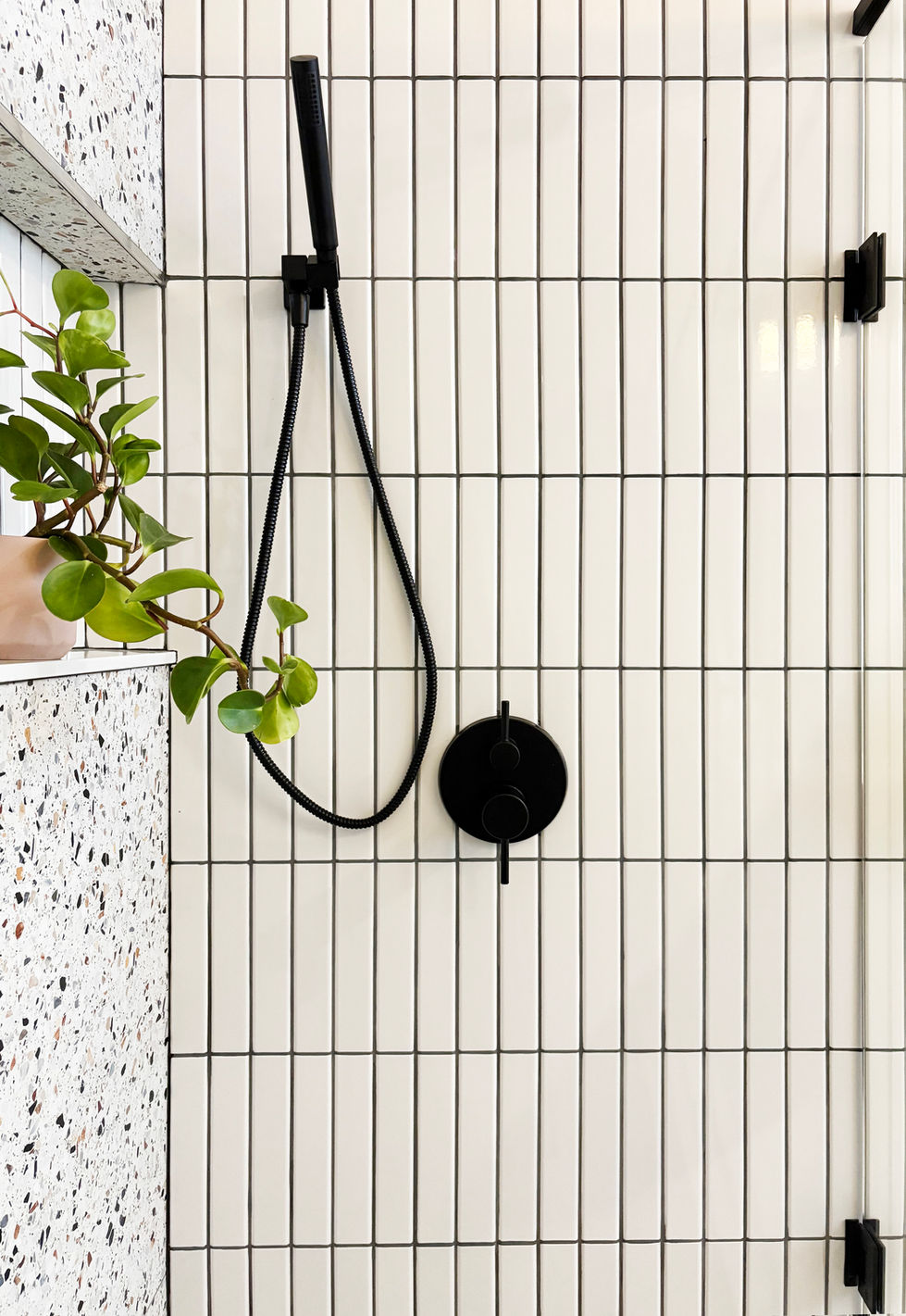 Vertical white tile shower wall with black handheld shower fixture and terrazzo detail.