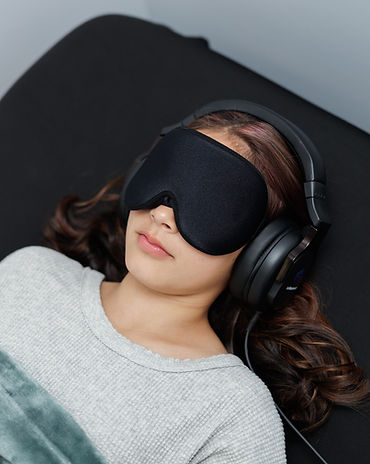Woman lying on black bed, wearing headphones, black eye mask, gray long sleeve, and covered with turqoise fuzzy blanket.