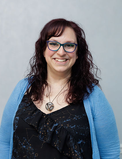 Headshot of woman smiling at camera. She's wearing blue purple ombre glasses, has dark red curly hair, with a blue cardigan and black top with a necklace.