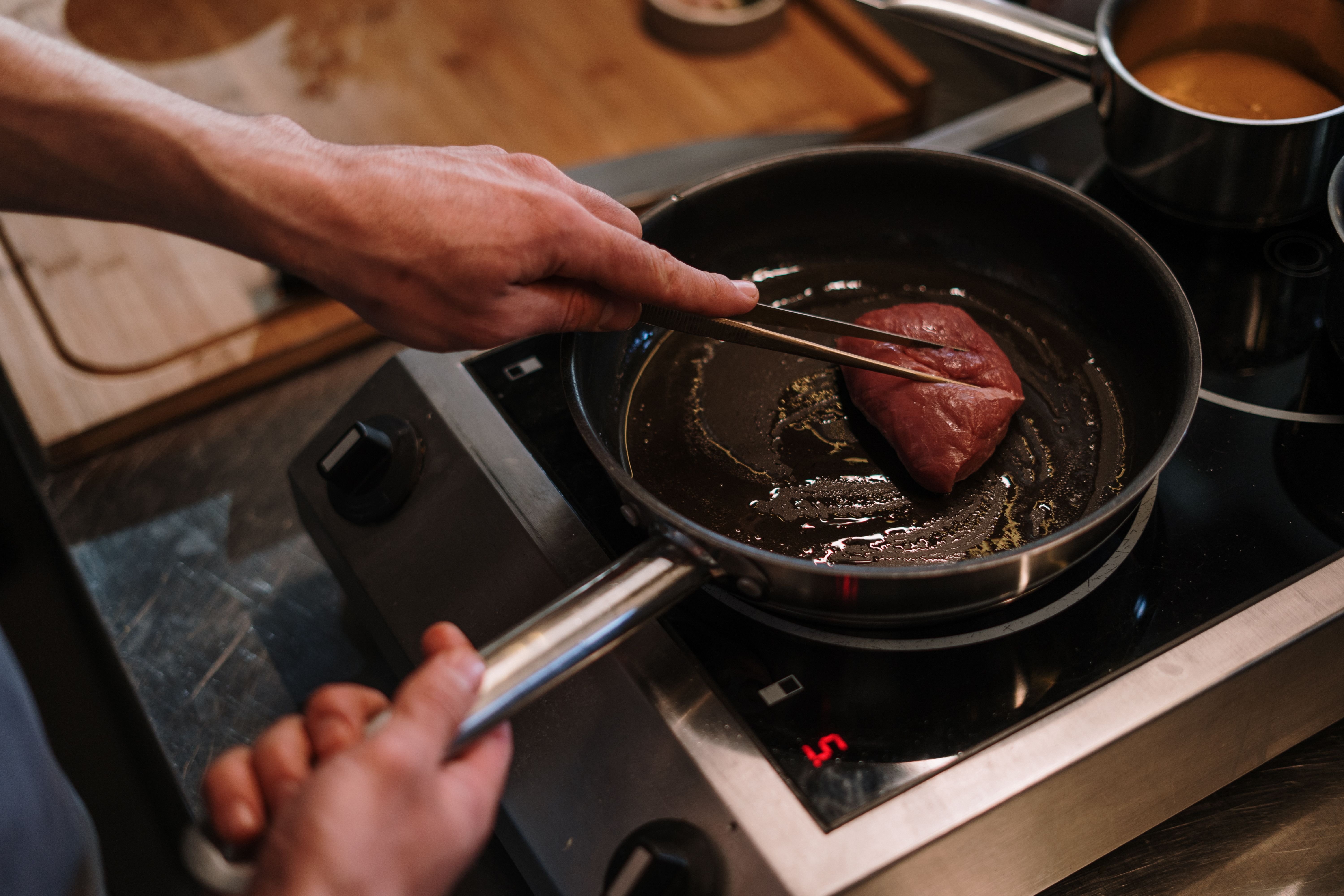 A chef searing a steak with tongs in a skillet in a modern professional kitchen