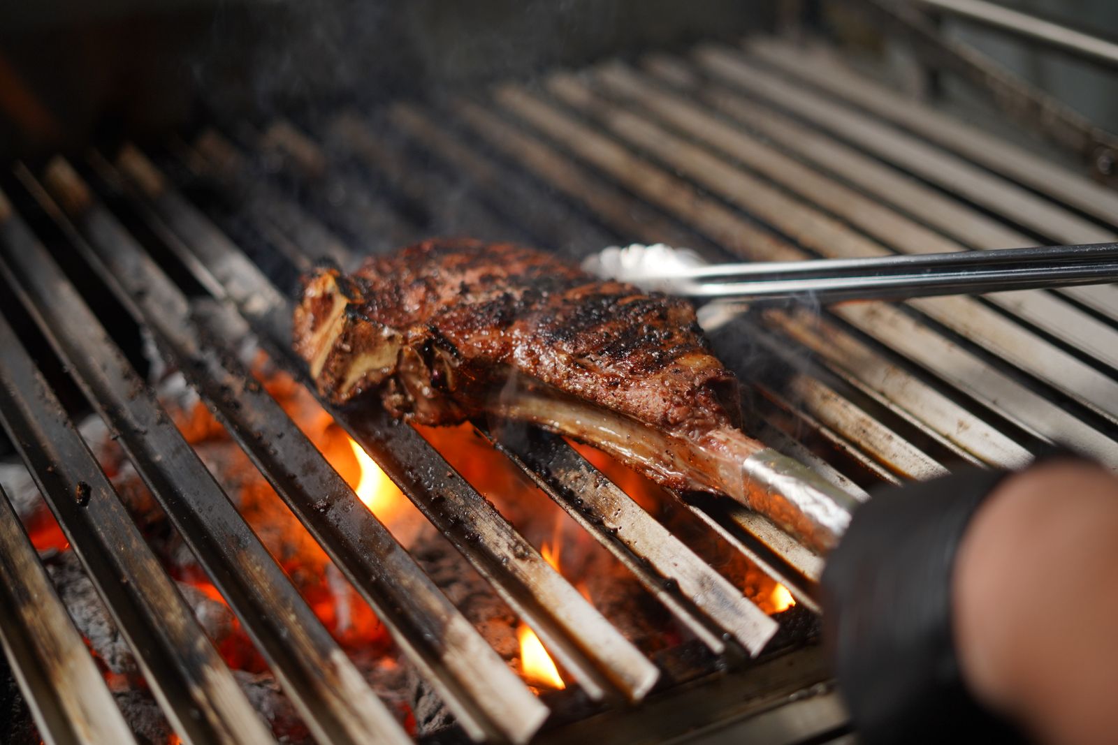 Tomahawk steak turned with tongs over flames on a grill