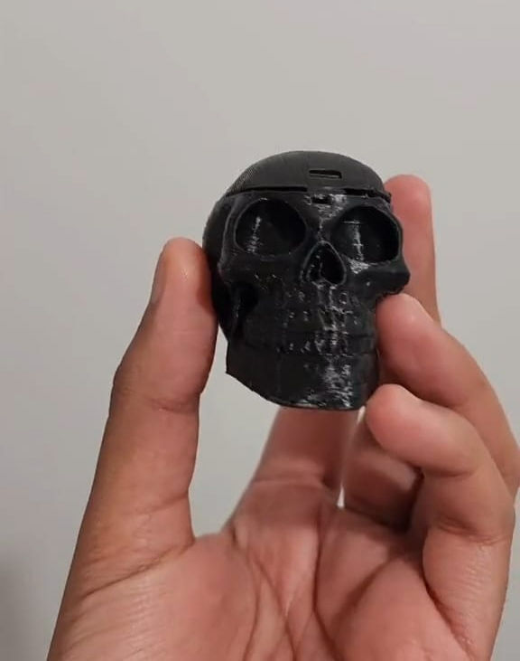 Mano sosteniendo una pequeña calavera negra impresa en 3D, con detalles definidos en los ojos, nariz y dientes. El fondo es liso y claro, lo que resalta la figura del cráneo.