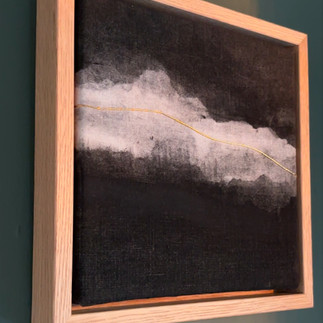 Minimalist abstract art piece featuring a soft gray cloud-like form against a dark background, with a thin golden line cutting through, displayed in a light wooden floating frame against a deep teal wall, showcasing contemporary monochromatic artwork with subtle metallic accent