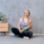 Elena Amani yoga instructor sitting cross-legged holding a colourful yoga mat, smiling, wearing grey top and black leggings, posture and Scenar therapy expert at Yoga for Posture Canberra.