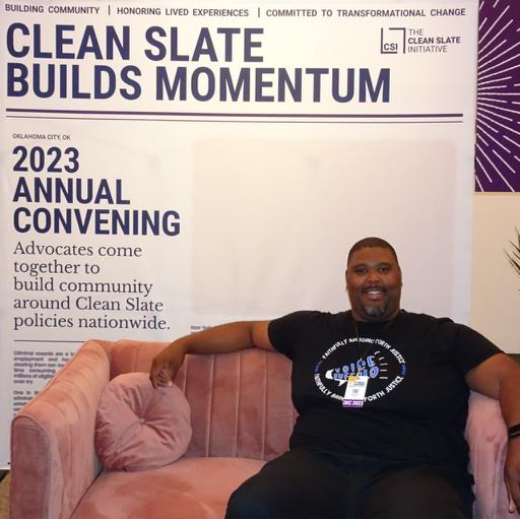 Black masculine presenting person sitting on couch in front of sign that reads Clean Slate Builds Momentum 2023 Annual Convening