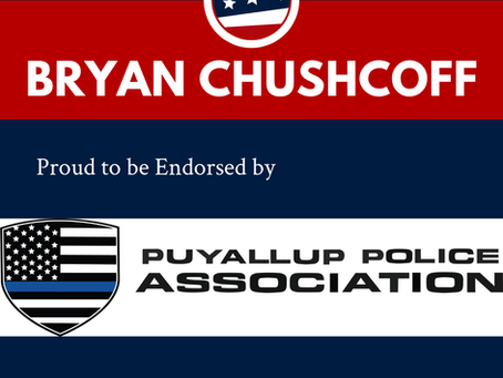 Puyallup Police Association logo featuring a black and white US Flag on a badge with a thin blue line announcing the endorsement of Bryan Chushcoff for Pierce County Prosecutor 2026