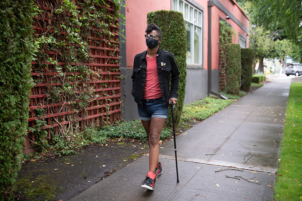 Black man with a black cane wearing a masking walking on a sidewalk