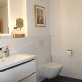 Modernes Badezimmer mit beleuchtetem Kosmetikspiegel, Waschtisch und im Spiegel sichtbarer Infrarotkabine in der FeWo MeerNah Grömitz | Whg. 3.
