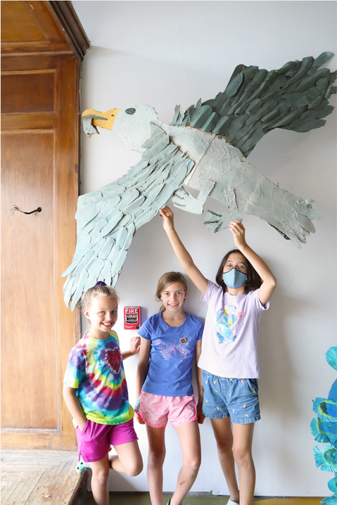 Children smiling beneath a large handmade bird sculpture displayed on a classroom wall