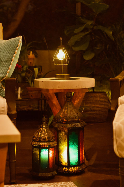 Covered lanai with tan concrete table top with backlit geode and pine wood base, lit Moroccan lanterns in the foreground, and plants in the background