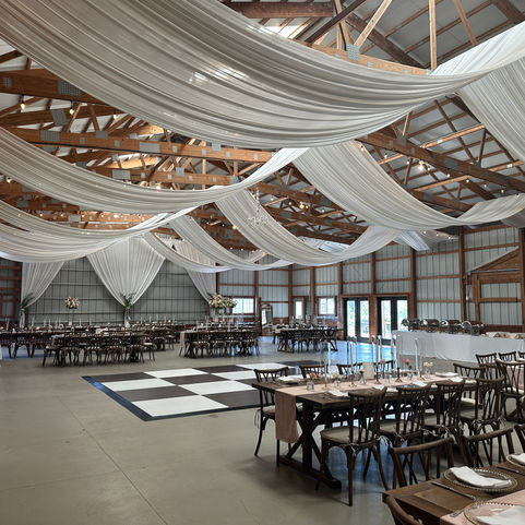barn wedding reception with straight open style white ceiling draping with bulb lighting bulb rope strands chiavari chairs with dark roast harvest tables 
