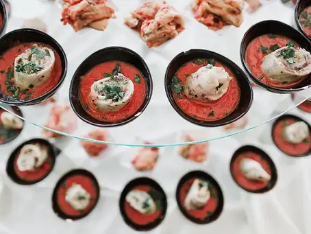 Photography by Drimage Creative studio by Simona Turano Fotografa. Assortment of appetizers in black bowls on a glass stand, with red sauce and a garnish of aromatic herbs on a white background.