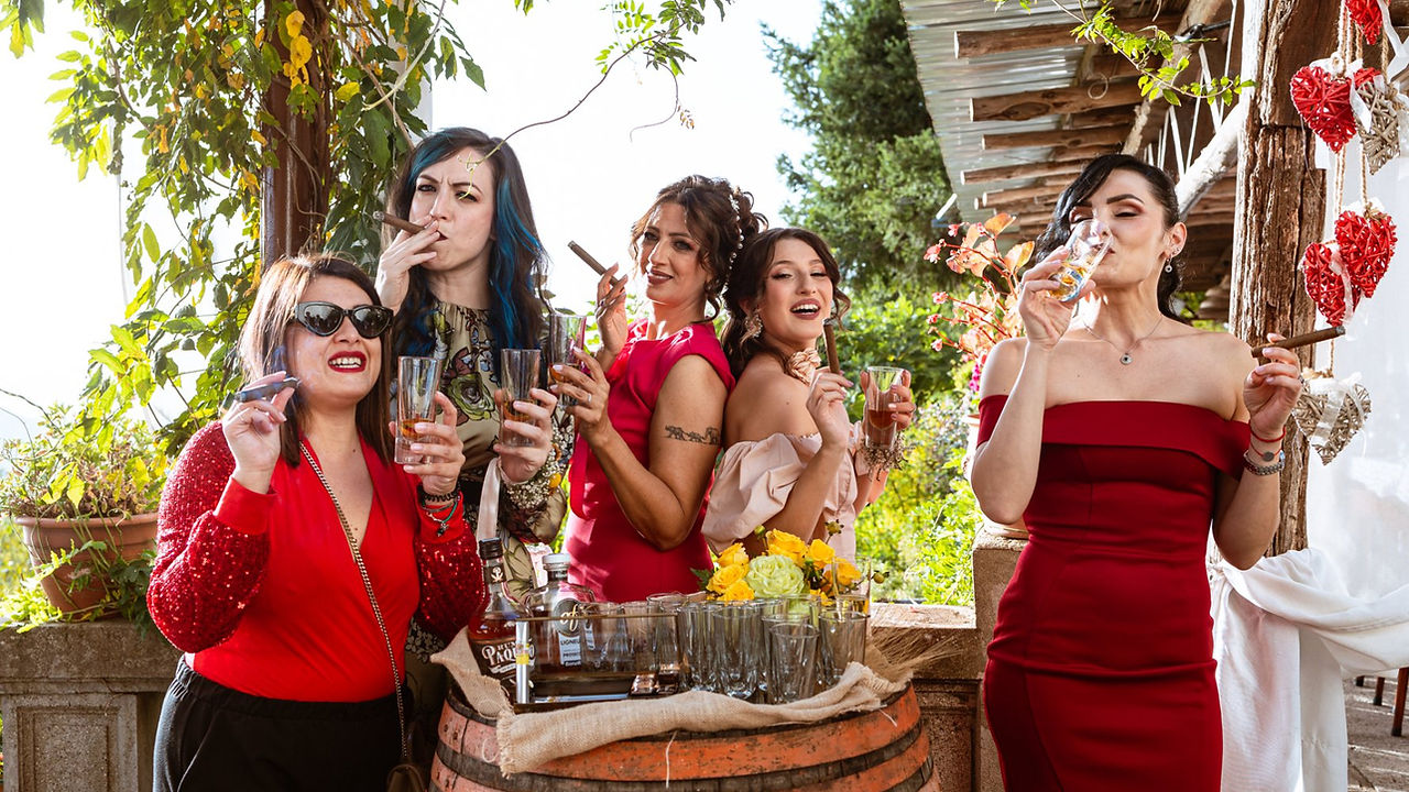 Cinque donne brindano con sigari e vino, risate felici, abiti rossi e festeggiamenti.